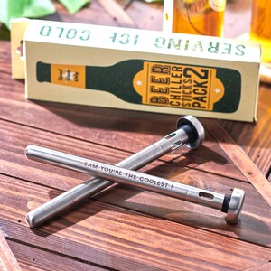Pode incluir: Dois palitos de refrigerar cerveja de a&ccedil;o inoxid&aacute;vel com uma ponta de borracha preta. Os palitos est&atilde;o cruzados um sobre o outro. O palito superior tem o texto "SAM YOU'RE THE COOLEST!" gravado. A embalagem dos palitos de refrigerar cerveja est&aacute; no fundo. A embalagem &eacute; verde e amarela e diz "SERVING ICE COLD" e "BEER CHILLER STICKS PACK 2".