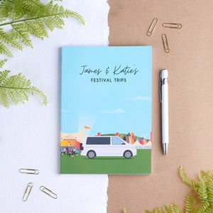 May include: A blue notebook with the text "James & Katie's Festival Trips" on the front cover. The notebook features a white van driving through a festival scene.