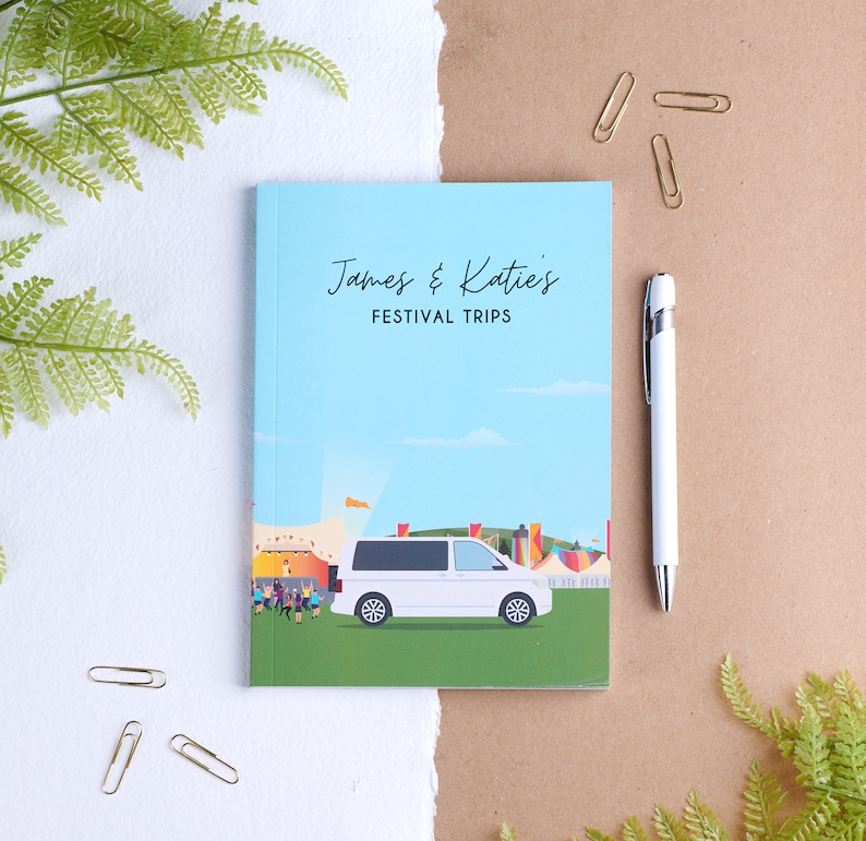 May include: A blue notebook with the text "James & Katie's Festival Trips" on the front cover. The notebook features a white van driving through a festival scene.