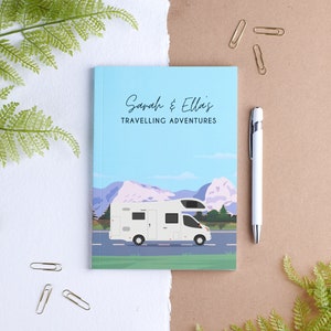 May include: A blue and green travel journal with the text "Sarah & Ella's Travelling Adventures" on the front cover. The journal features a white camper van driving on a road with mountains in the background.