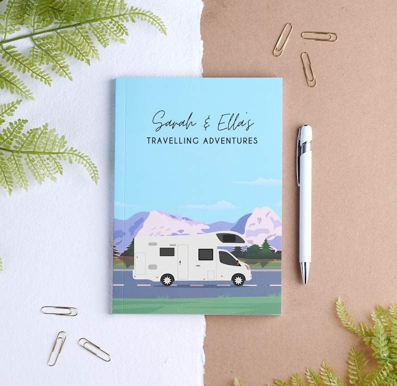 May include: A blue and green travel journal with the text "Sarah & Ella's Travelling Adventures" on the front cover. The journal features a white camper van driving on a road with mountains in the background.