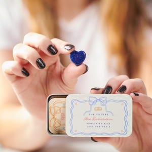 Puede incluir: Un pequeño objeto azul en forma de corazón se sostiene sobre una lata rectangular abierta. La lata tiene una tapa blanca con un borde azul y el texto "THE FUTURE Mrs. Richardson SOMETHING BLUE JUST FOR YOU X". Dentro de la lata hay un objeto rectangular pequeño con un diseño floral.