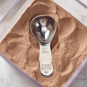 Puede incluir: Una cuchara medidora de acero inoxidable con el texto "THOMAS I'M WHEY INTO YOU" y un diseño de corazón, en un recipiente con polvo marrón. La cuchara está marcada con "2 T 30ml".