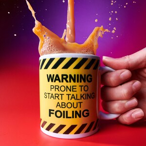 May include: A white ceramic mug with a yellow and black warning design. The mug reads "WARNING PRONE TO START TALKING ABOUT FOILING." A liquid is splashing out of the mug. The mug is held by a hand.