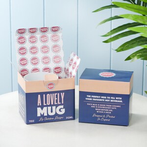Puede incluir: Una taza de cer&aacute;mica blanca con el texto "A Lovely Mug" en letras rosa y azul. La taza est&aacute; empaquetada en una caja rosa y azul con el texto "11oz" y "312ml" impreso en el lateral. La caja tambi&eacute;n incluye el texto "By Oakdene Designs".