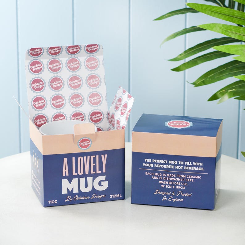 Puede incluir: Una taza de cer&aacute;mica blanca con el texto "A Lovely Mug" en letras rosa y azul. La taza est&aacute; empaquetada en una caja rosa y azul con el texto "11oz" y "312ml" impreso en el lateral. La caja tambi&eacute;n incluye el texto "By Oakdene Designs".
