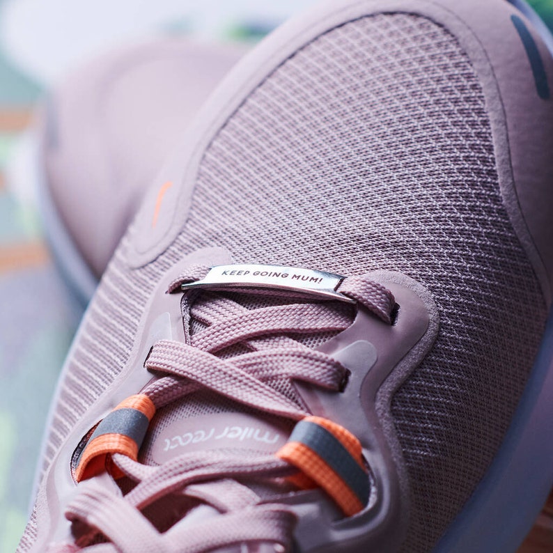 K&ouml;nnte beinhalten: Nahaufnahme eines Paares rosa Laufschuhe mit einem silbernen Metalletikett mit der Aufschrift "KEEP GOING MUMI!" Die Schuhe haben orangefarbene Akzente an den Schn&uuml;rsenkeln und ein strukturiertes Gewebe.