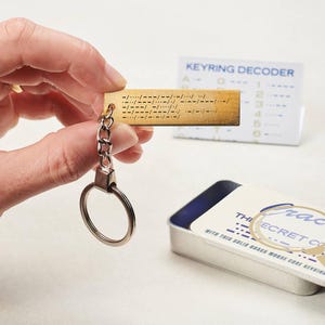 May include: A brass Morse code keyring with a chain and key ring is held in front of a keyring decoder card. The keyring has Morse code symbols engraved on it. A small metal tin with text is in the background.