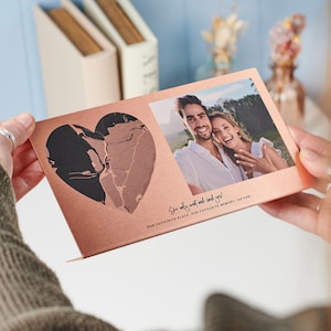 May include: A rose gold metal card with a black and rose gold heart design. The card has a photo of a couple smiling at the camera. The text on the card reads "She only went and said yes! Our favourite place, our favourite memory (so far)".