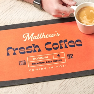 Könnte beinhalten: Orangefarbene und schwarze Barmatte mit dem Text "Matthew's Fresh Coffee" in einer Retro-Schrift. Der Text "ESTD 1992" befindet sich ebenfalls auf der Matte, sowie der Text "ENJOYED IN BRIGHTON, EAST SUSSEX" und "COMING IN HOT!"