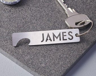 May include: A silver bottle opener with the name "JAMES" cut out, a key, and a bottle cap are on a gray surface. The bottle opener has a key ring hole and a curved end for opening bottles. The key is silver and has a standard key shape.