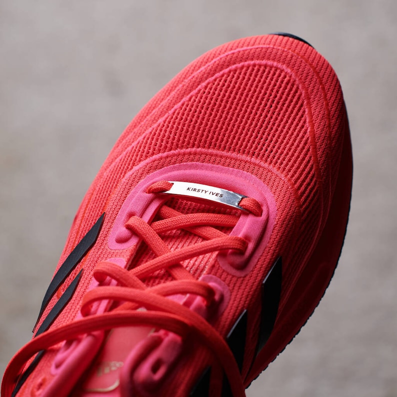 K&ouml;nnte beinhalten: Nahaufnahme eines roten Laufschuhs mit schwarzen Akzenten. Der Schuh hat ein silbernes Schild mit dem Text "Kirsty Ives" an den Schn&uuml;rsenkeln.