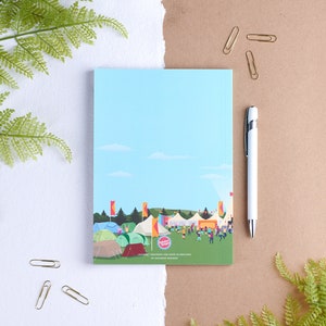 May include: A blue notebook with a colorful illustration of a campsite with tents, trees, and people. The text "Festival Friends" is printed on the cover. The notebook is designed and made in England by Garden Studio.