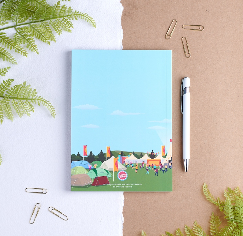 May include: A blue notebook with a colorful illustration of a campsite with tents, trees, and people. The text "Festival Friends" is printed on the cover. The notebook is designed and made in England by Garden Studio.
