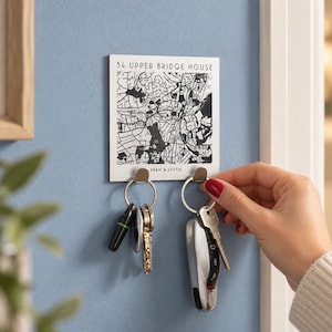 May include: A square wall-mounted key holder featuring a black and white street map print, the text 34 UPPER BRIDGE HOUSE, and the names ADAM & LOTTIE. The white board has two silver metal hooks, each holding a silver keyring with various keys attached.