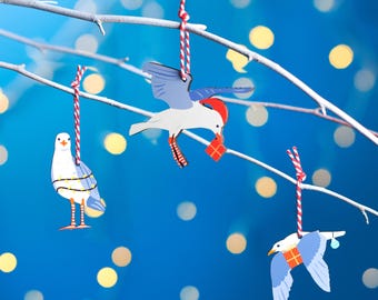 以下が含まれることがあります： 枝にぶら下がっている木製の鳥のオーナメントが3つ。鳥は白く塗られており、青い羽根と赤いアクセントが付いています。1羽の鳥が小さな赤いギフトボックスを持っています。オーナメントは、ぼやけた白い光が点滅する青い背景に映えています。