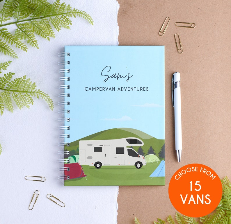 May include: A blue and green notebook with a white camper van illustration and the text "Sam's Campervan Adventures". The notebook has a spiral binding and is on a white surface. There is a white pen and gold paper clips on the surface.