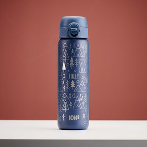 Könnte beinhalten: Eine marineblaue, isolierte Wasserflasche mit einem Waldmotiv. Sie zeigt eine weiße Umrissillustration von Bäumen, Bergen, einem Bären und dem Namen "FINLEY". Der Markenname "ION8" ist unten aufgedruckt.