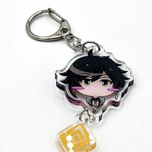 May include: A clear acrylic keychain featuring a chibi-style character with black hair, amber eyes, and a small dangling charm shaped like a yellow square with a geometric symbol. The keychain includes a silver-toned metal lobster clasp ring.