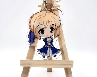 May include: A clear acrylic keychain featuring a chibi-style anime character with blonde hair and green eyes. The character wears a blue dress with white trim and a blue bow. The keychain is attached to a silver metal keyring.