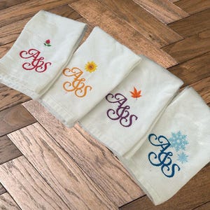Pode incluir: Um conjunto de toalhas de mão brancas com tecido de waffle, cada uma bordada com as iniciais "AJS" em uma escrita decorativa. Cada toalha apresenta um motivo sazonal diferente: uma rosa, um girassol, uma folha de bordo e um floco de neve.