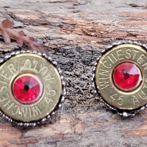 May include: A pair of earrings made from repurposed .45 caliber bullet casings with red rhinestones in the center. The casings are marked "WINCHESTER 45 AUTO".