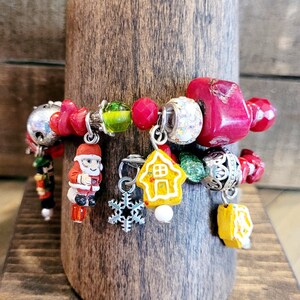 May include: A festive Christmas charm bracelet with red, silver, and yellow accents. The bracelet features Santa Claus, gingerbread house, and snowflake charms. The bracelet is displayed on a wooden stand.