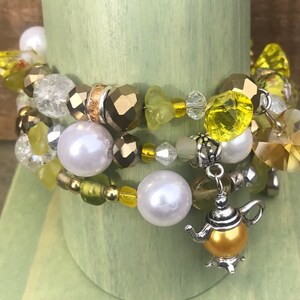 May include: A stack of three beaded bracelets with a variety of beads in yellow, gold, clear, and white. The bracelets include faceted beads, pearls, and a teapot charm. The bracelets are displayed on a light green cylindrical stand.