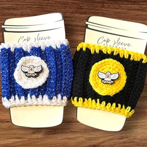 May include: Two handmade crochet cup sleeves. One is blue and white with a silver thread, the other is black and yellow. Each has a circular emblem with a bird design. The text "Cup sleeve" is visible.