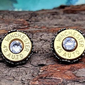 May include: A pair of gold-colored bullet casing earrings with a clear rhinestone in the center. The casings are engraved with "Winchester 45 Auto".