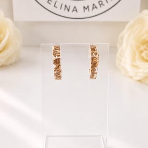 May include: A pair of hoop earrings featuring a clear resin base embedded with small, metallic gold-toned rectangular pieces. The earrings are displayed on a clear acrylic stand in front of a white card with the text TELINA MARIE.