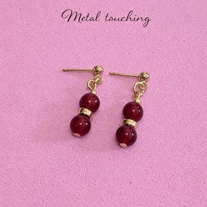 May include: A pair of gold-colored earrings with a post back. Each earring features two dark red, round beads separated by a gold-colored spacer. The text "Metal touching" is above the earrings.