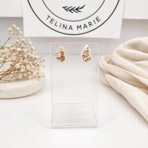 May include: A pair of teardrop-shaped earrings featuring a white base with embedded gold leaf flakes. The earrings are displayed on a clear acrylic stand in front of a white card with the text Handmade TELINA MARIE printed in black ink.