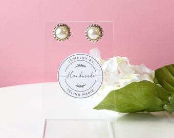 May include: A pair of pearl earrings on a clear display stand. The earrings are gold-toned with a small pearl in the center. The stand has a sticker that says "Jewelry by Handmade Telina Marie".