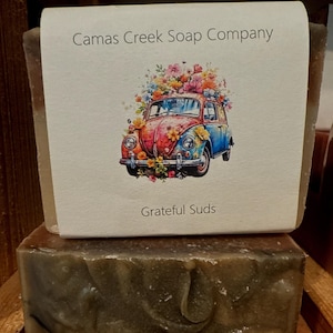 May include: A bar of soap with a brown and green color. The soap is wrapped in a white paper label with a colorful illustration of a vintage car with flowers on top. The label reads "Camas Creek Soap Company" and "Grateful Suds".