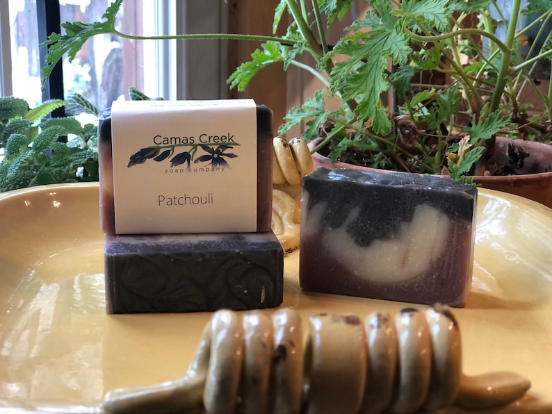 Pu&ograve; includere: Tre saponette con l'etichetta "Camas Creek Soap Company". La saponetta superiore &egrave; etichettata "Patchouli". I saponi sono scuri con un design a strati. Un dosatore di miele &egrave; in primo piano su un piatto giallo.
