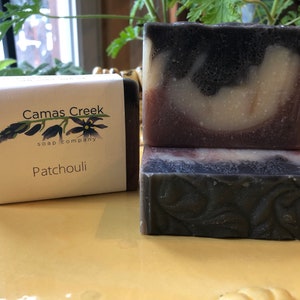 Pu&ograve; includere: Tre saponette Patchouli di Camas Creek. Una saponetta &egrave; avvolta con un'etichetta. Il sapone ha un colore scuro con vortici pi&ugrave; chiari e un motivo floreale. L'etichetta recita "Camas Creek soap company" e "Patchouli".