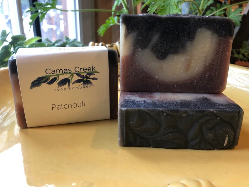 Pu&ograve; includere: Tre saponette Patchouli di Camas Creek. Una saponetta &egrave; avvolta con un'etichetta. Il sapone ha un colore scuro con vortici pi&ugrave; chiari e un motivo floreale. L'etichetta recita "Camas Creek soap company" e "Patchouli".