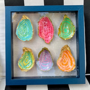 Puede incluir: Una obra de arte enmarcada con seis conchas de ostra pintadas. Cada concha es de un color diferente, incluyendo azul, rosa y naranja, con bordes dorados y patrones de puntos y espirales blancos. El marco es azul liso.