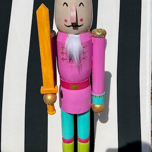 Puede incluir: Una caprichosa figura de cascanueces de madera pintada a mano. Presenta una chaqueta rosa, pantalones turquesa y botas verde lima. La corona es verde y rosa, y sostiene una espada dorada. La base es rosa brillante.