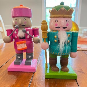 May include: Two wooden nutcracker figurines, one in pink with a drum and the other in green with a scepter. Both have white beards and smiling faces.