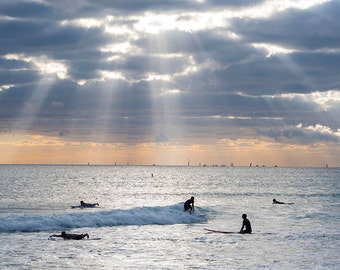 May include: Silhouettes of people surfing in the ocean on a cloudy day with sunbeams breaking through the clouds.