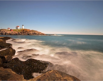May include: A white lighthouse with a red roof sits on a rocky island with a blue sky and ocean in the background. The ocean waves are blurred, creating a soft, dreamy effect.