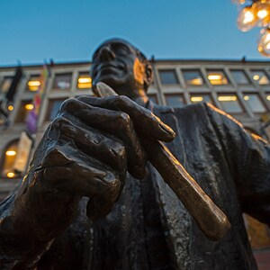 May include: Bronze statue of a man holding a baton, set against a backdrop of buildings and a twilight sky. The statue's details are highlighted by the warm glow of streetlights and building lights, creating a contrast of light and shadow.