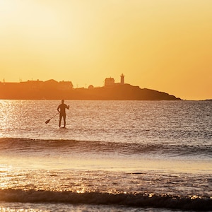 May include: Silhouette of a person paddleboarding in the ocean at sunrise with a lighthouse in the distance.