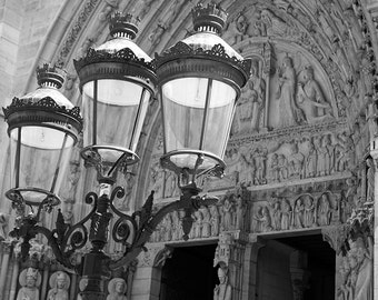 May include: A black metal lamp post with three ornate lampshades in front of a stone archway with intricate carvings. The lampshades are made of glass and have a clear finish.