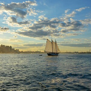 May include: A white sailboat with sails billowing in the wind, sailing on a calm blue body of water with a city skyline in the background. The sun is setting behind the city, casting a golden glow on the water.