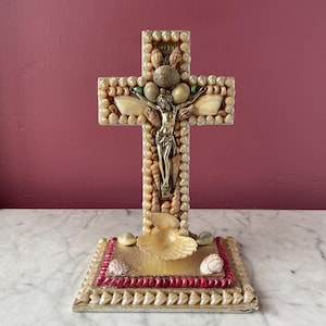 Puede incluir: Una cruz decorativa hecha de conchas marinas, con una figura de latón de Jesucristo. La cruz está adornada con varias conchas y piedras, y se apoya sobre una base rectangular con un borde rojo, también decorada con conchas.