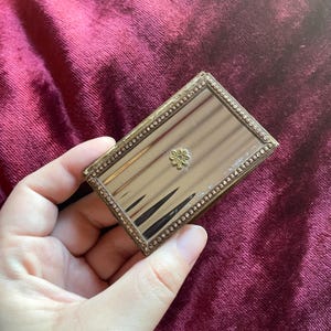 May include: Rectangular antique compact mirror with a gold-toned frame and a small floral embellishment. The mirror's surface has a reflective quality, and the frame is adorned with a beaded border. The compact is held against a burgundy background.