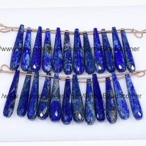 Könnte beinhalten: Eine Strähne aus 30 tropfenförmigen Lapislazuli-Edelsteinkugeln mit goldfarbenen Drahtschlaufen. Die Perlen sind etwa 2,5 cm lang und haben eine dunkelblaue Farbe mit weißen und goldenen Flecken. Die Perlen sind auf einem goldfarbenen Draht aufgereiht.